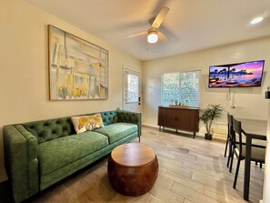 A living room with a green sofa, a round wooden coffee table, a ceiling fan, a TV, and a window with blinds.