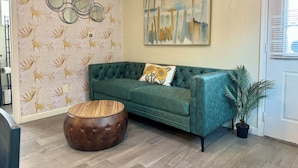 A green sofa with a sunflower pillow, a round wooden ottoman, a wall with floral wallpaper, and a painting above the sofa. - Restaurants, Cafes & Close to Downtown & Coronado (San Diego)