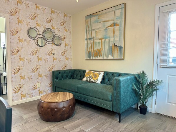 A green sofa with a sunflower pillow, a round wooden ottoman, a wall with floral wallpaper, and a painting above the sofa.