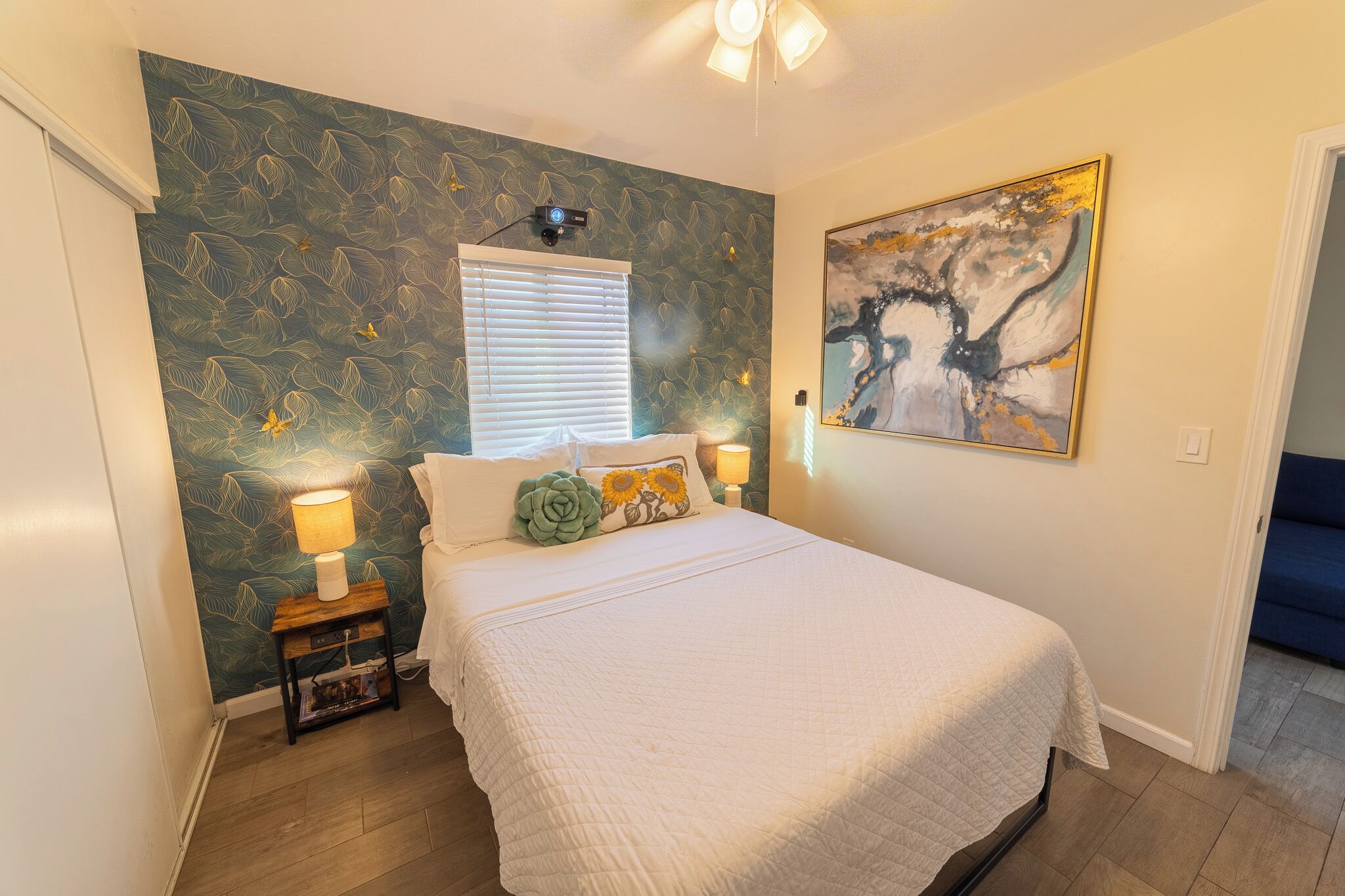 Een slaapkamer met een bed, een nachtkastje, een lamp, een raam met jaloezieën en een schilderij aan de muur.