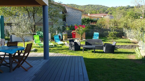 Een terras met buitenmeubilair, een overdekt gedeelte en uitzicht op een huis en omliggend groen.