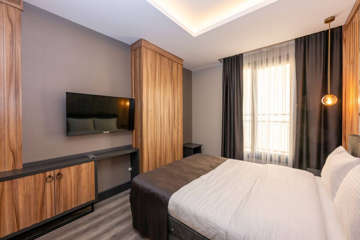 A modern hotel room with a large bed, wooden wardrobe, built-in TV, and a large window with curtains.