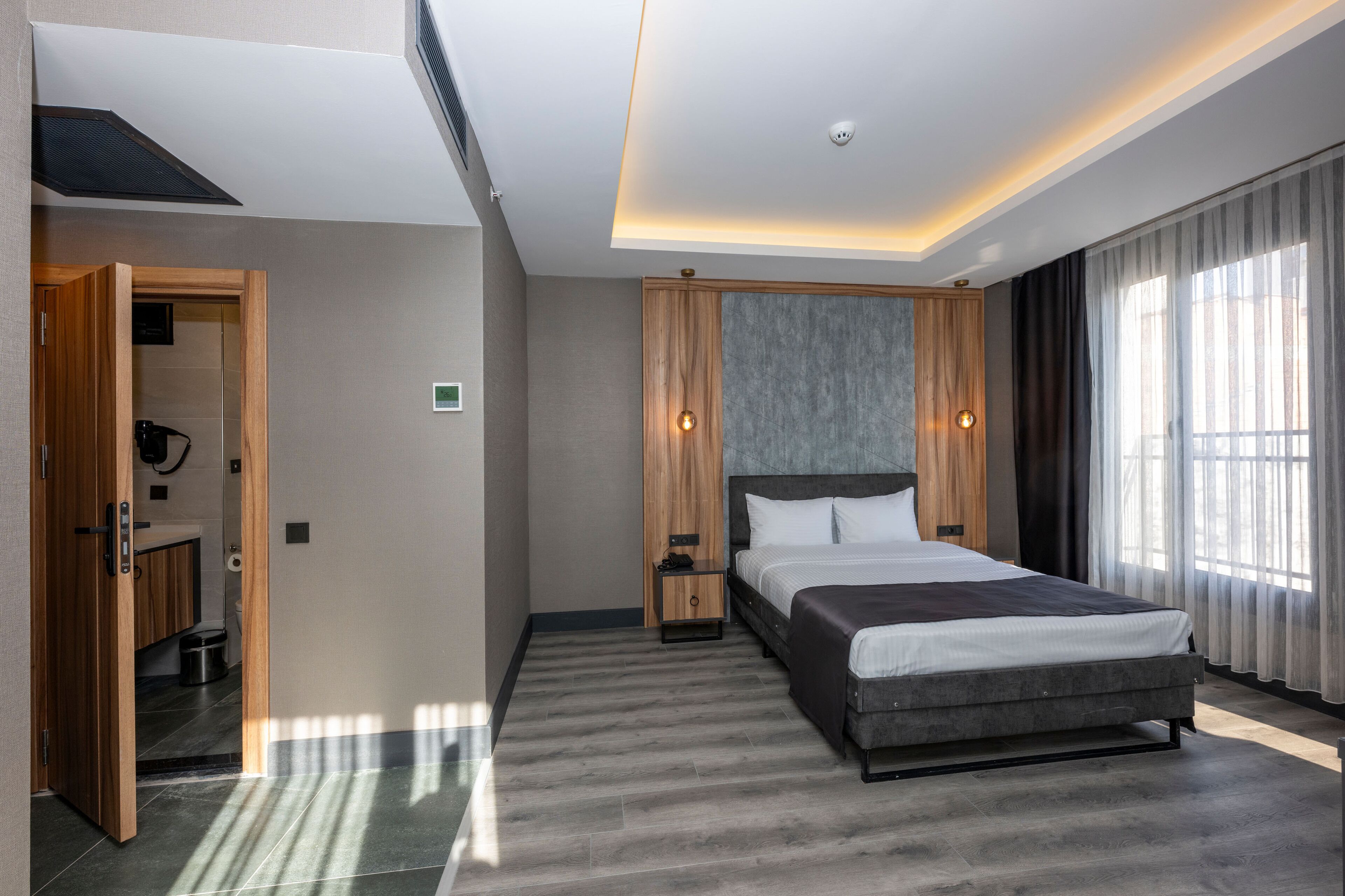 Une chambre d’hôtel moderne avec un grand lit, une armoire en bois, une petite table de chevet et une salle de bain visible à travers une porte ouverte.