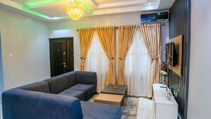 A modern living room with a sectional sofa, a small side table, a flat-screen TV mounted on the wall, and a chandelier.