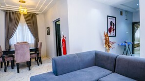 A modern living room with a grey sofa, a dining table with chairs, a chandelier, a fire extinguisher, and a framed picture on the wall.