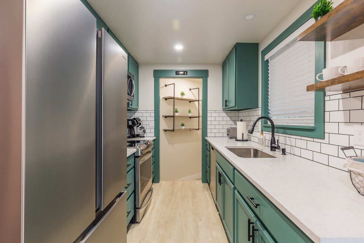 A modern kitchen with teal cabinets, a stainless steel refrigerator, a built-in oven, and a white countertop with a sink and faucet.