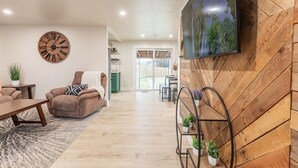 A modern living room with a wooden herringbone wall, a large flat-screen TV, a round wooden clock, a brown sofa with a patterned pillow, a coffee table, and a small metal plant stand. - Centrally Located Family Home, Backyard w Firepit! (Meridian)