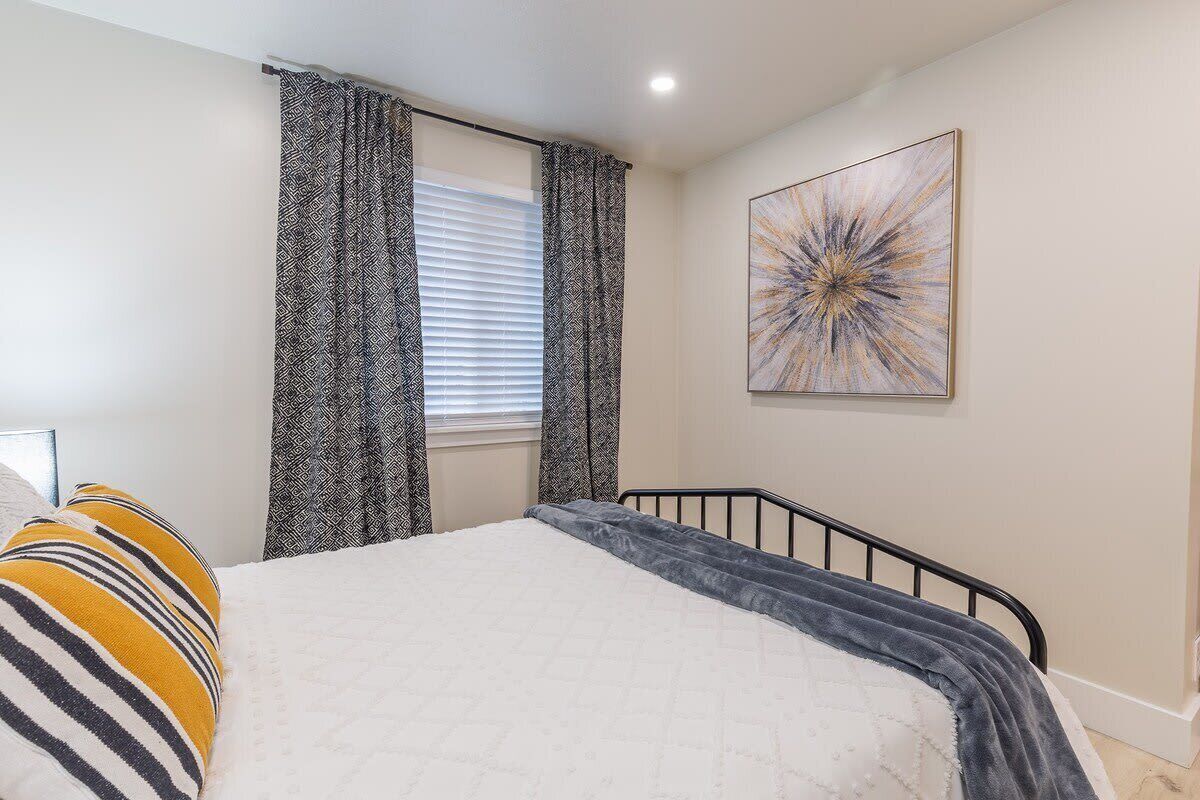 A bedroom with a bed, patterned curtains, a window with blinds, and a wall-mounted abstract art piece.