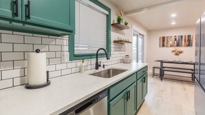 A modern kitchen with teal cabinets, a white countertop, a stainless steel sink, and a dishwasher. There is a window with blinds, a small table with a vase, and a painting on the wall. - Centrally Located Family Home, Backyard w Firepit! (Meridian)