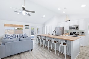 Una cocina moderna con armarios blancos, una isla central, taburetes de bar, un ventilador de techo y un ventanal grande.