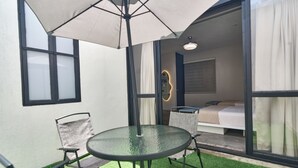 Una habitación con puerta de cristal que da a un balcón, una cama, una mesa redonda de cristal con sillas y un paraguas grande.