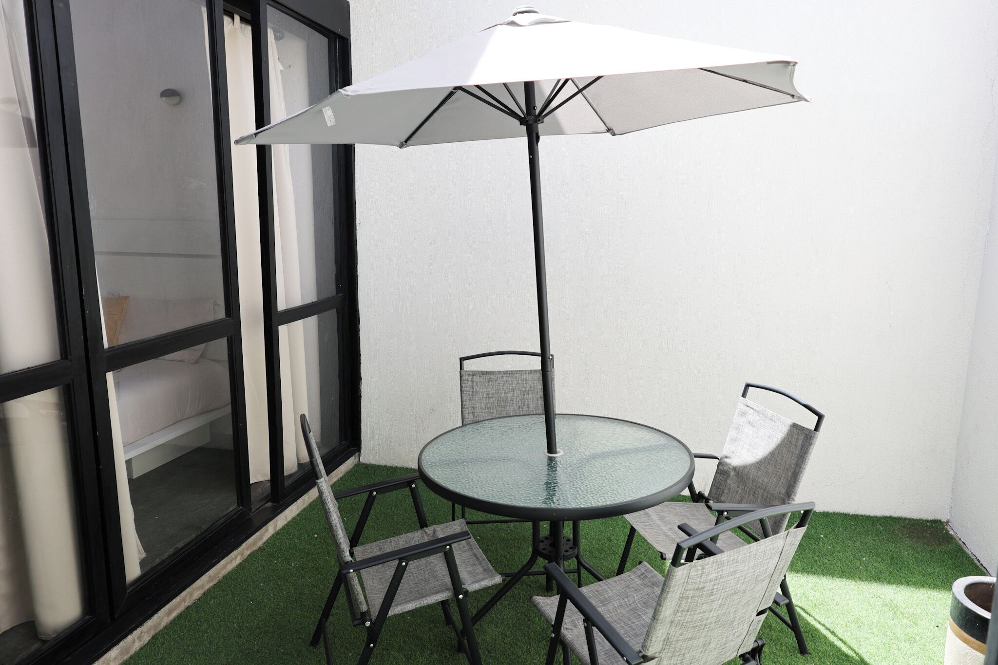 A glass table with four chairs and a white umbrella in a small outdoor area with artificial grass.