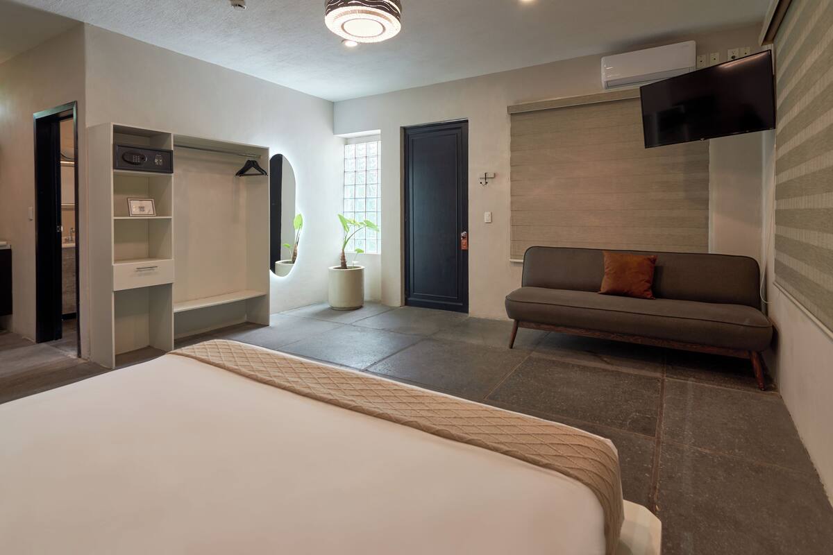 Una habitación de hotel con cama, sofá, vestidor, planta en maceta y televisor fijado a la pared.