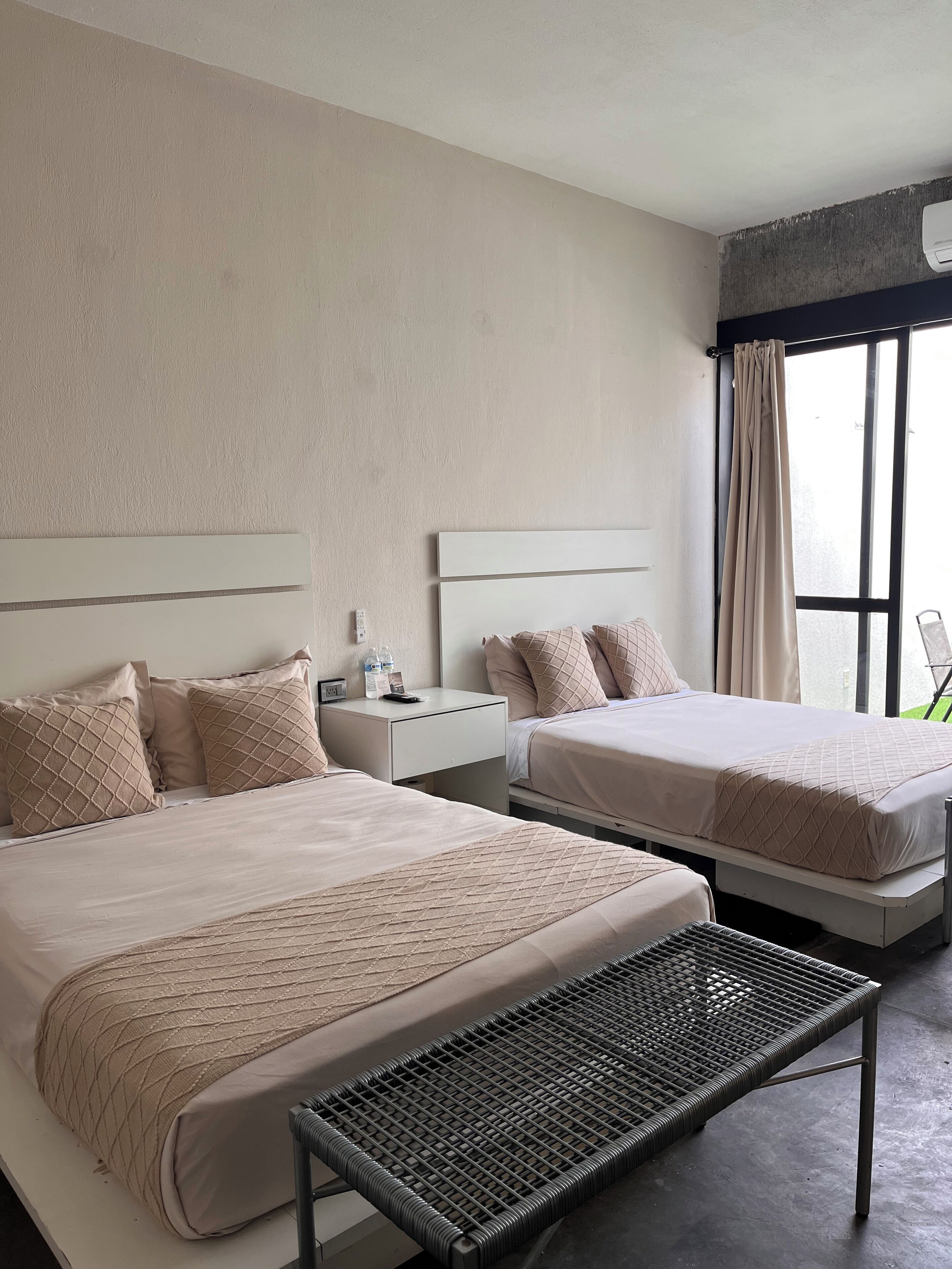 A hotel room with two beds, a small bedside table with a phone and water bottles, a bench, and a large window with curtains.