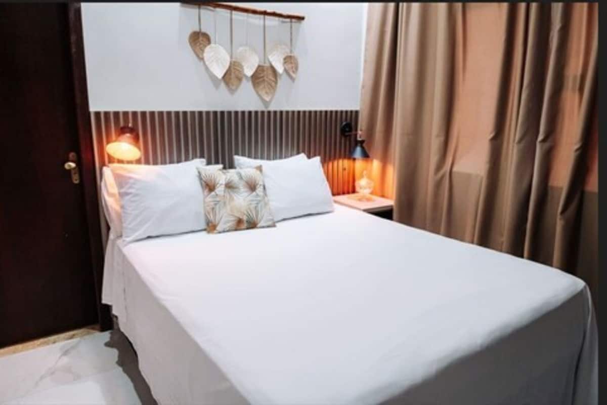 A neatly made bed with white linens and decorative pillows, a wall-mounted light fixture, and a curtain on the window.
