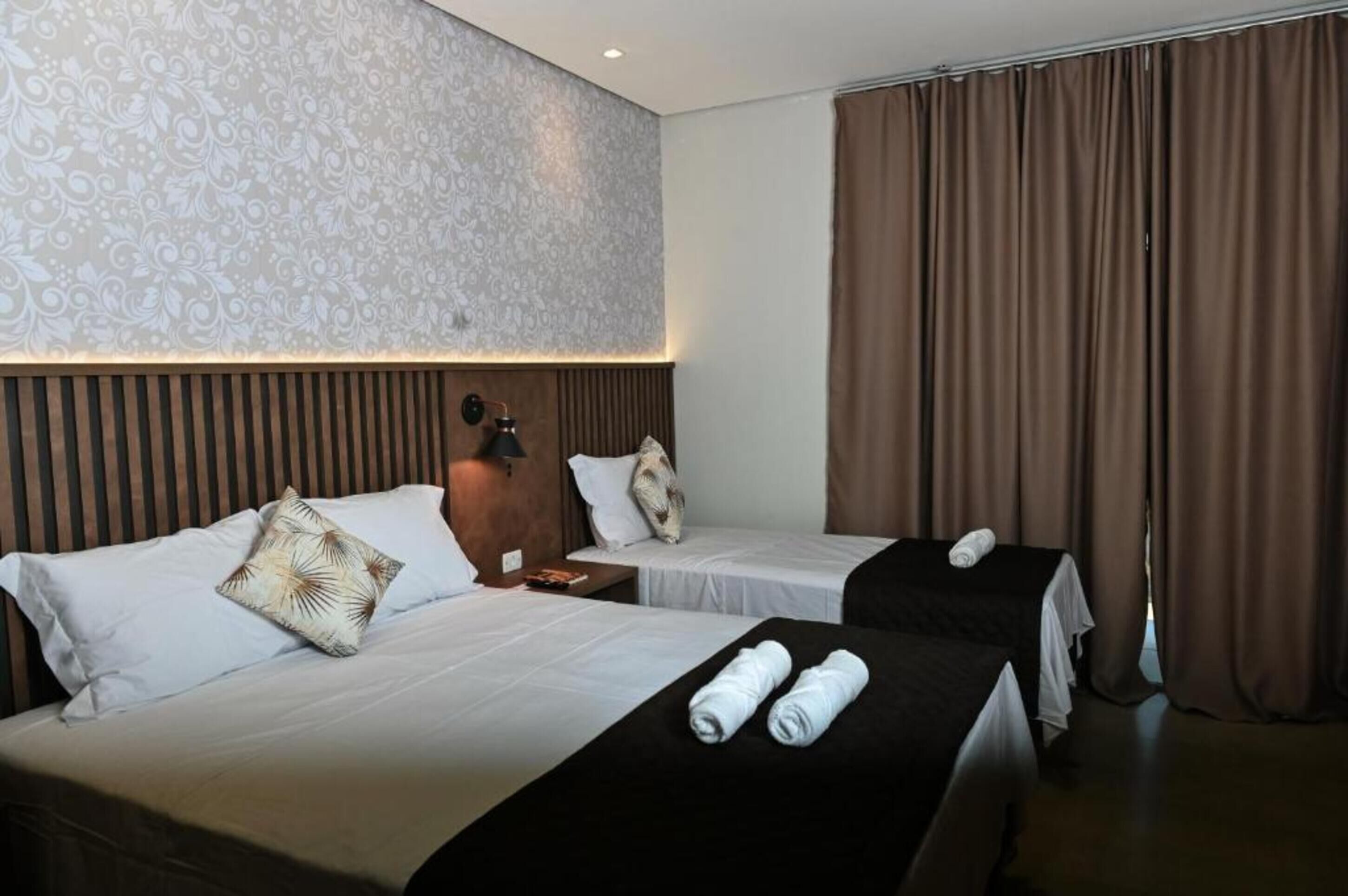 Habitación de hotel con dos camas, un papel tapiz estampado, cabecero de madera y un ventanal con cortinas.
