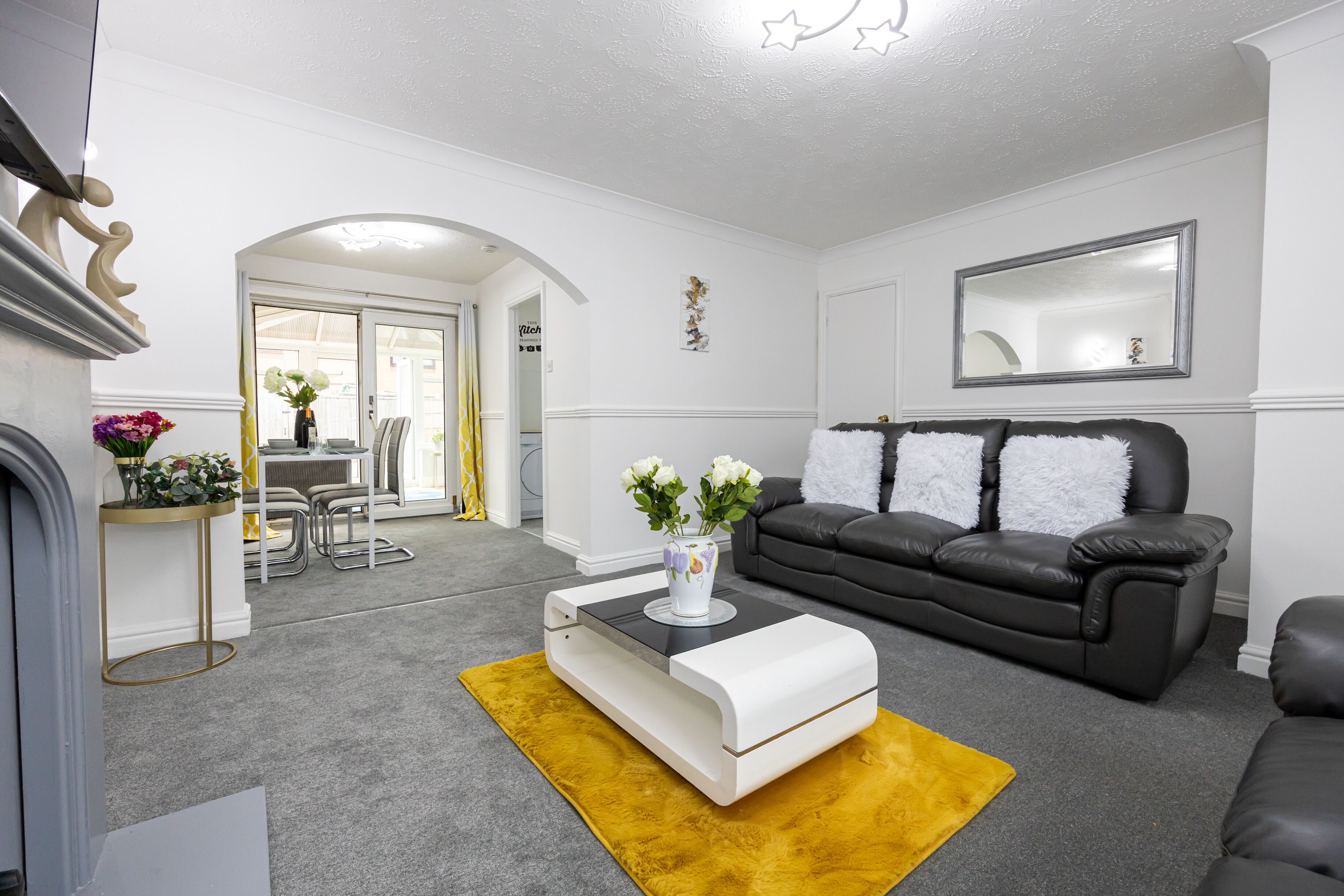 Ein modernes Wohnzimmer mit einem schwarzen Ledersofa, einem weißen Couchtisch und einem gelben Teppich. Im Hintergrund befindet sich ein Essbereich mit Tisch und Stühlen.