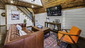 A cozy living room with a brown leather sofa, a wooden coffee table, an orange armchair, and a large TV mounted on the wall. - Little Woods- Luxury A-frame cabin |Firepit|Hottub (Broken Bow)