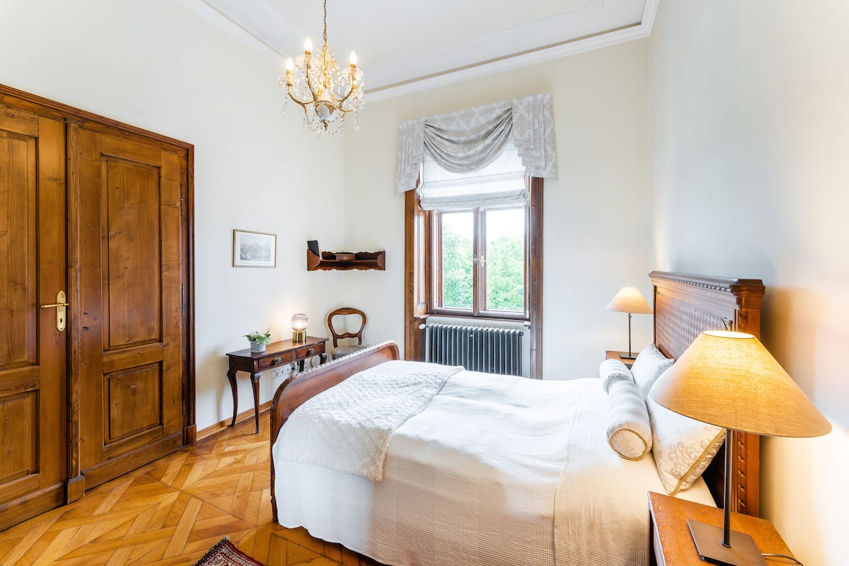 A bedroom with a large wooden wardrobe, a bed with a white bedspread, a bedside table with a lamp, and a window with a view of greenery.