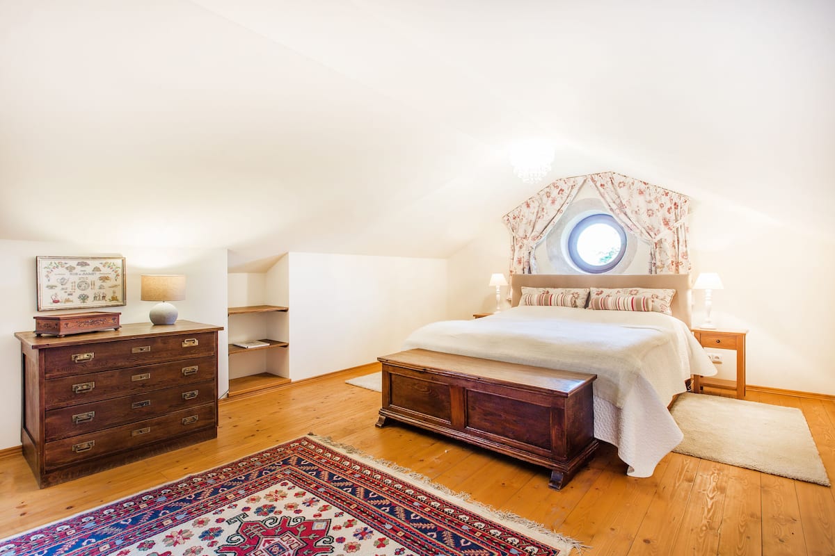 A bedroom with a wooden dresser, a large bed with a canopy, a round window, and a patterned rug.