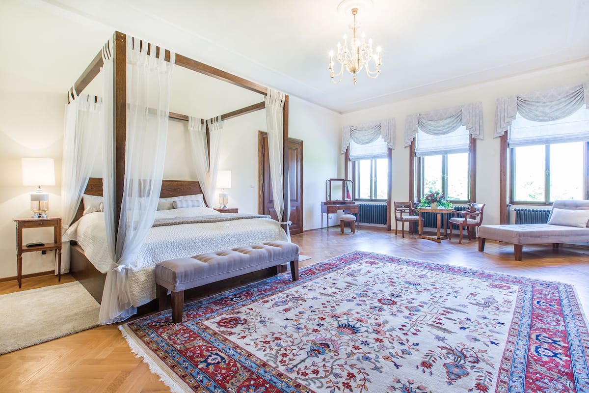 A spacious bedroom with a four-poster bed, a large ornate rug, and a chandelier.