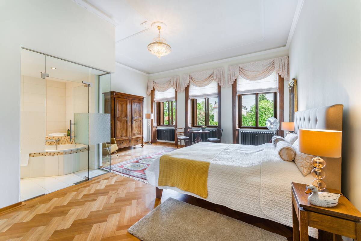 A bedroom with a large bed, a bedside table with a lamp, a window with curtains, a view of the outdoors, and a bathroom with a glass shower enclosure.
