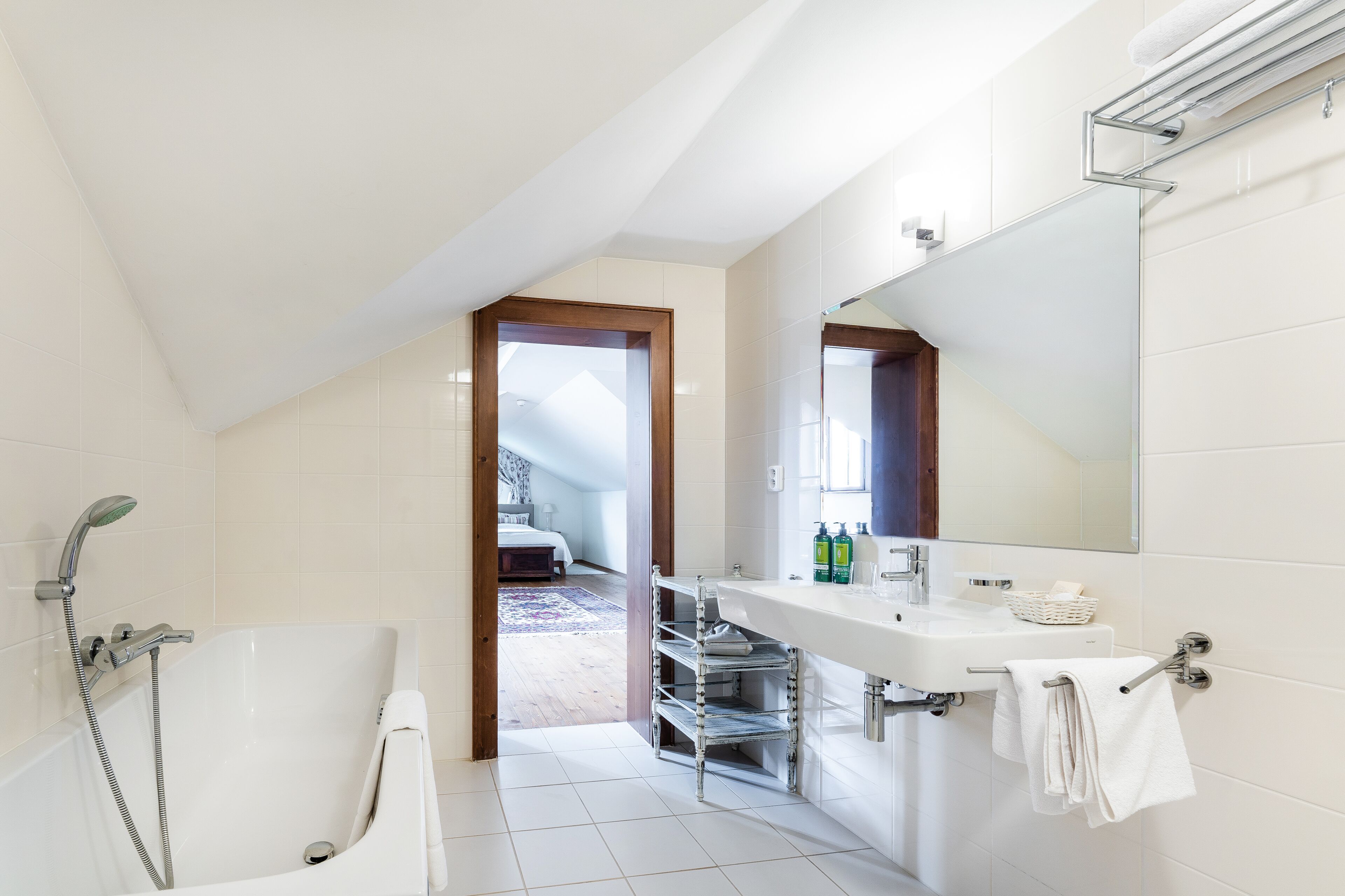 Een moderne badkamer met een bad, een wastafel en een douche. Boven de wastafel hangt een spiegel en er is uitzicht op de slaapkamer door een geopende deur.