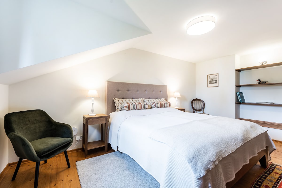 A bedroom with a bed, a chair, a nightstand, and a shelf.