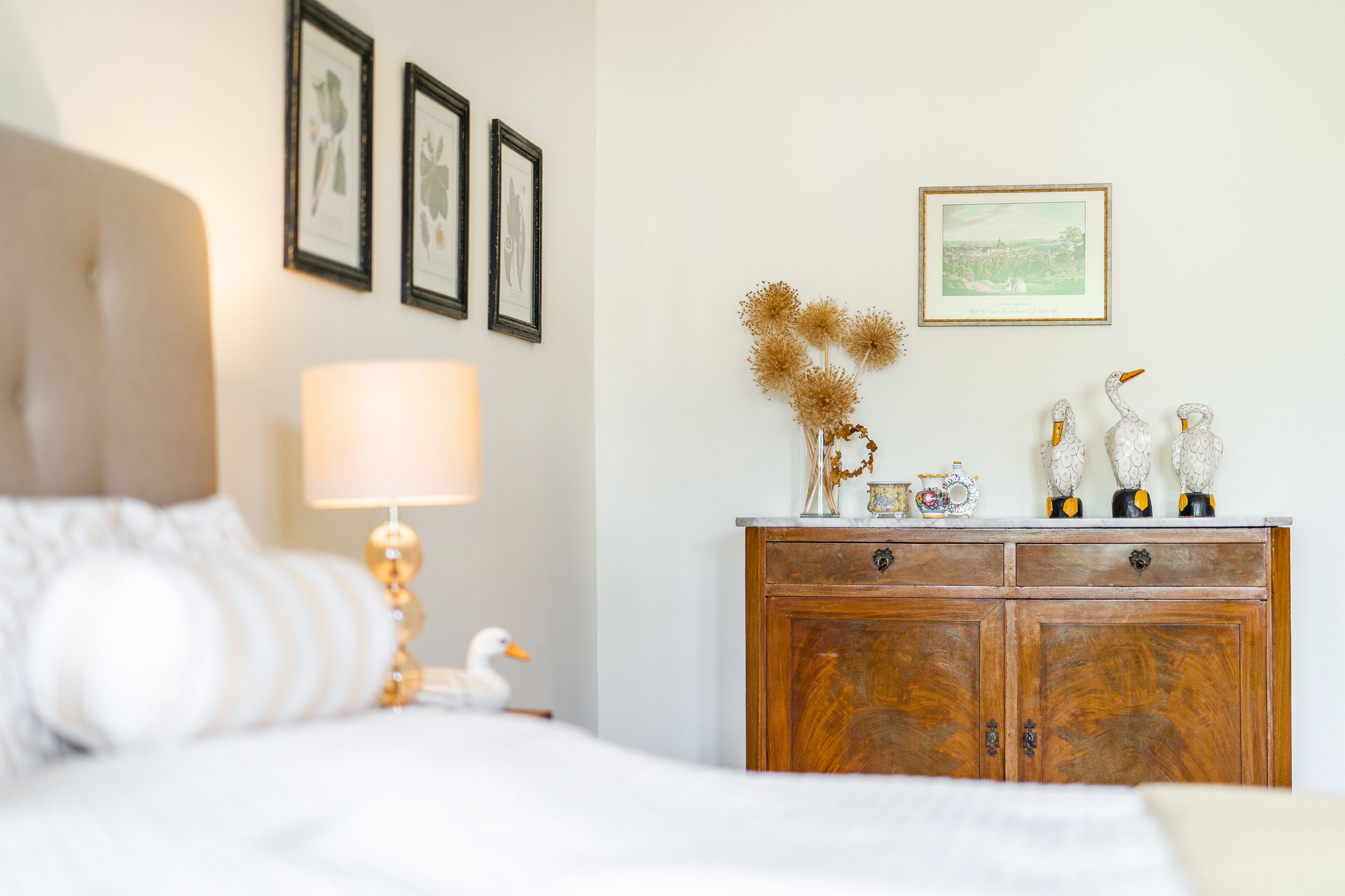 A bedroom with a bed, a nightstand with a lamp, a dresser with decorative items, and framed pictures on the wall.