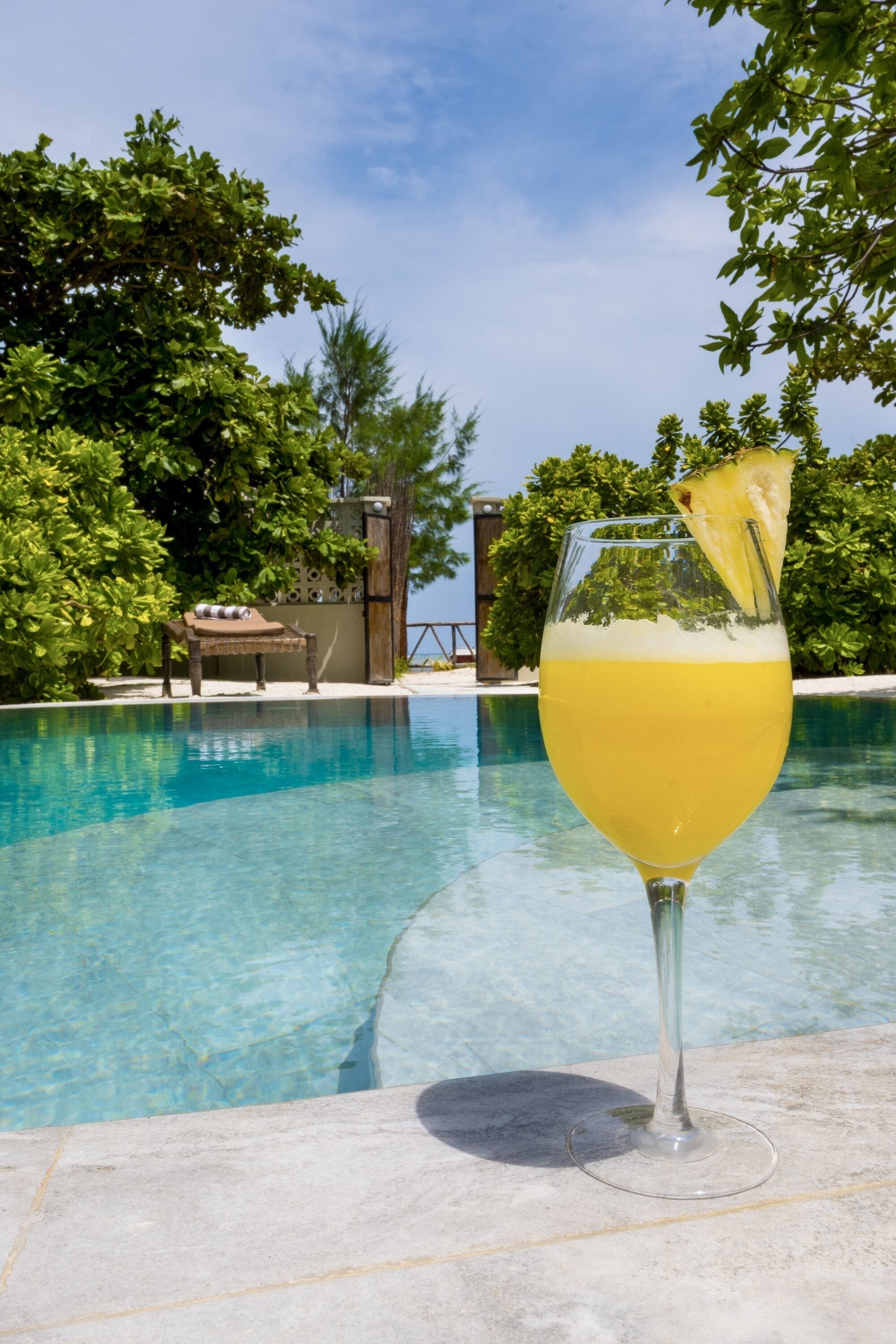 A glass of yellow drink with a pineapple garnish by a poolside.