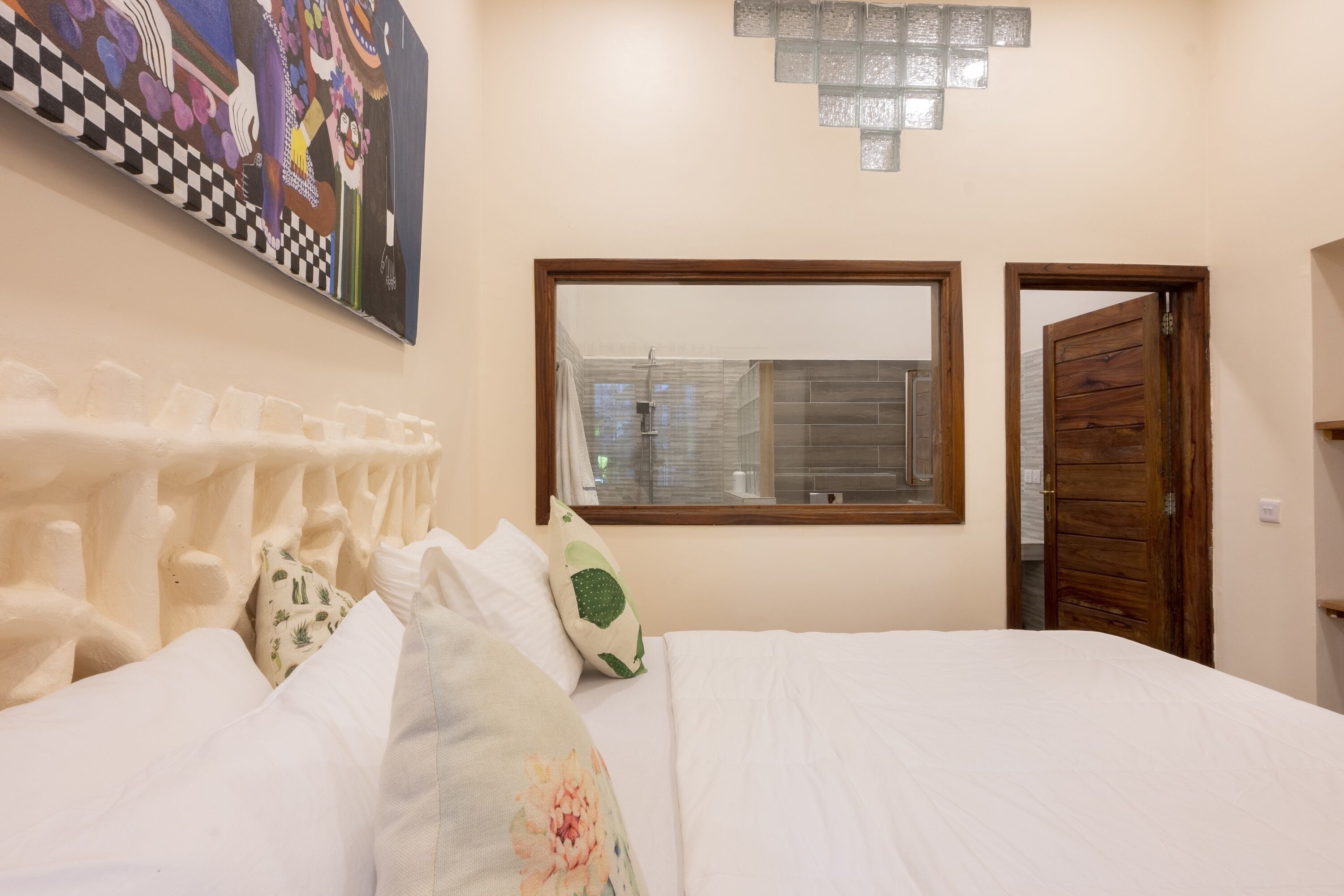 Une chambre à coucher avec un lit, des oreillers, une fenêtre, une porte et une œuvre d’art accrochée au mur.