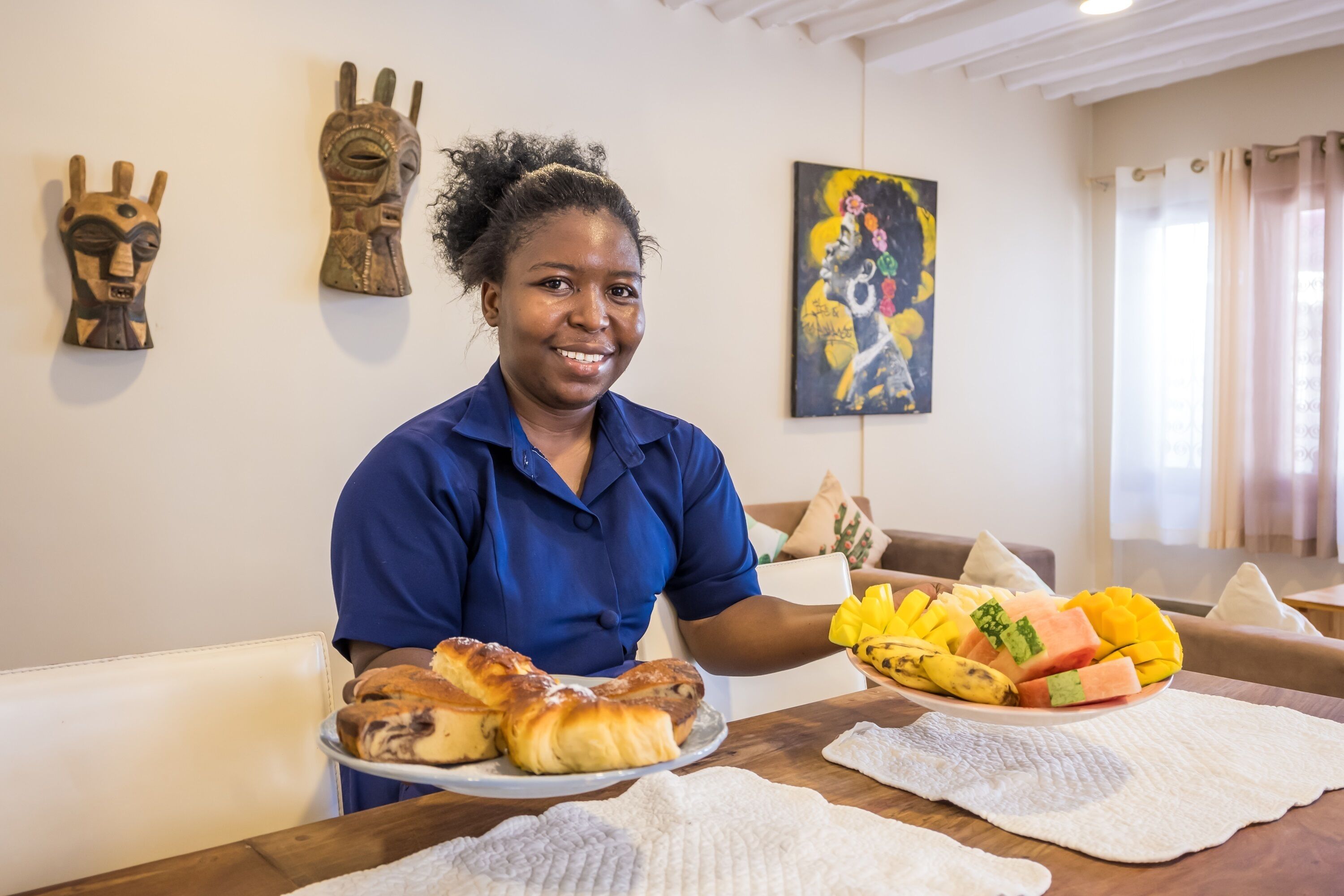 A person holding a plate of pastries and a bowl of fruit in a room with a white wall, a painting, and a couch.