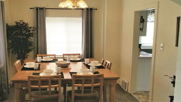 A dining room with a wooden table set for four, surrounded by chairs. There is a large window with curtains, a potted plant, and a chandelier. - Heart of the Falls! Cozy for 2, Spacious for 8, Close to everything! (Cuyahoga Falls)