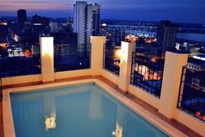 Una piscina en la azotea con vistas a la ciudad, rodeada de rascacielos y ventanas iluminadas.