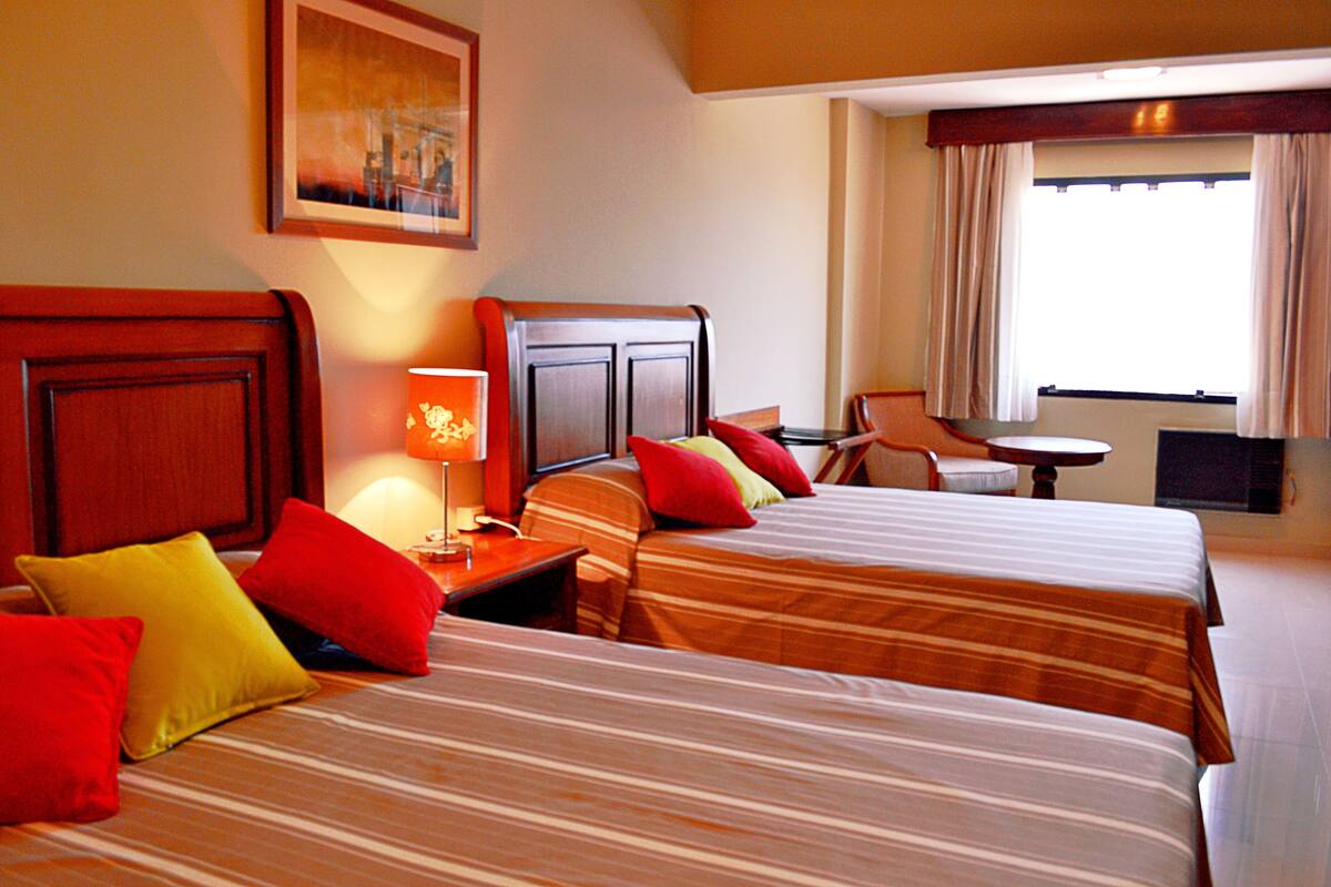 A hotel room with two beds, a nightstand, a lamp, a chair, and a window with curtains.