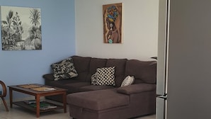A living room with a sofa, a coffee table, and a dining table. There are various fruits and vegetables on the table, including bananas, avocados, and a bowl of sweet potatoes.