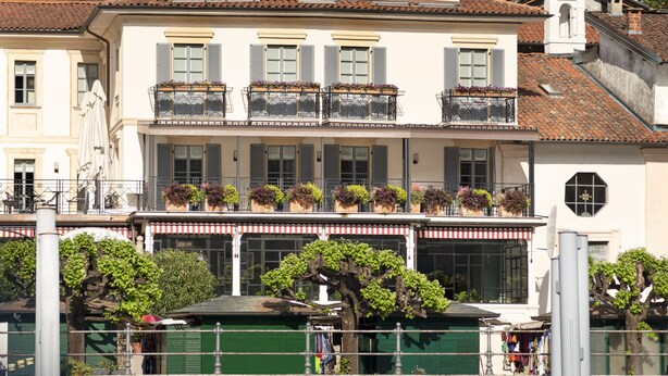 Een gebouw aan het water genaamd 'Albergo Delfino Ristorante' met balkons, plantenbakken en een stenen muur.