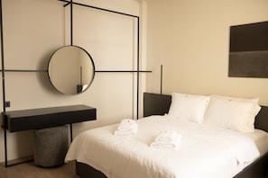 A modern bedroom with a bed, white linens, a round mirror, a black bedside table, and a gray ottoman. - Taskou Luxury Suites (Thessaloniki)