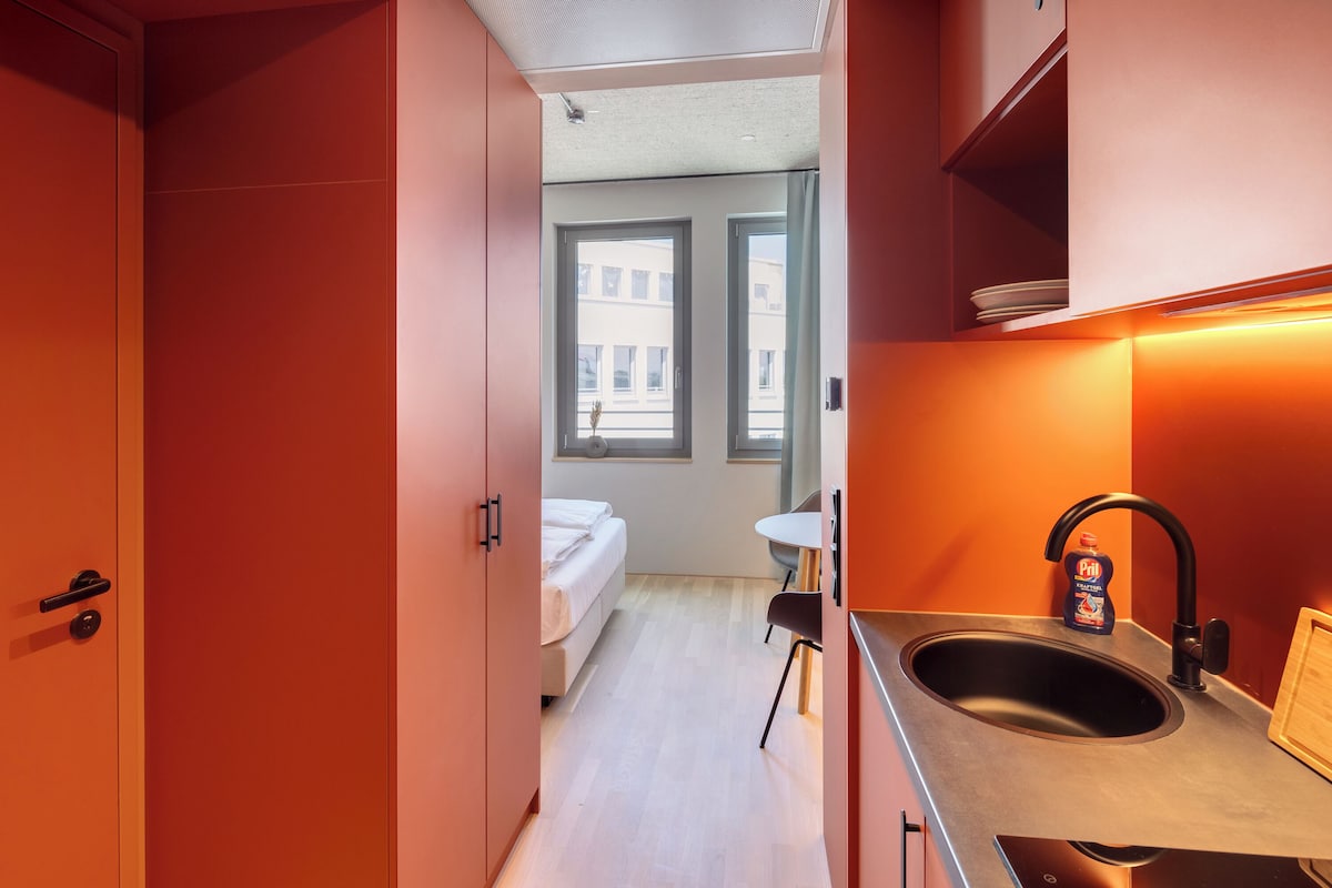 A compact hotel room with a kitchenette, a bed, and a view of the city through a window.