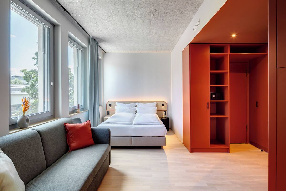 A modern bedroom with a bed, a grey sofa, a red pillow, a nightstand, a bedside table, and a wardrobe.