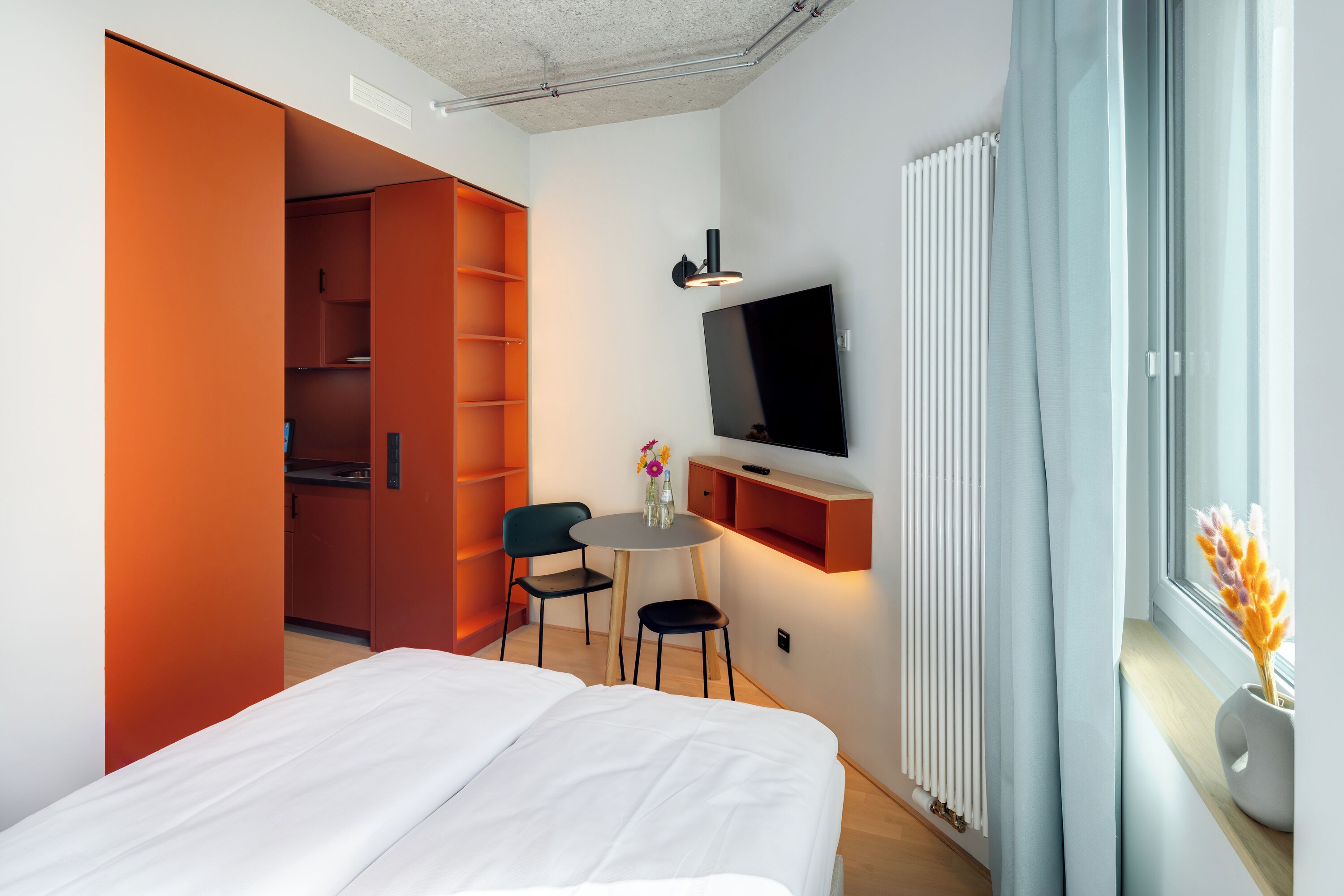 Quarto de hotel com cama, escrivaninha com cadeiras, televisão e janela com cortinas.
