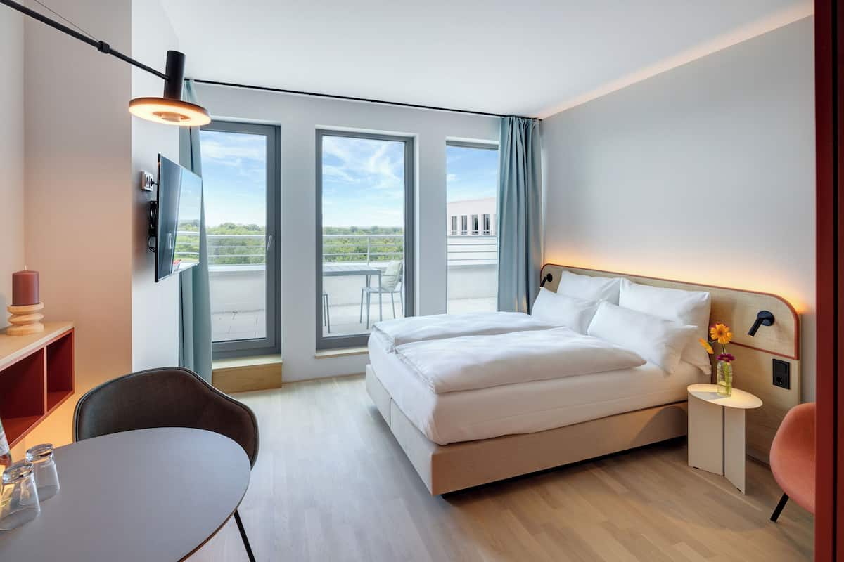 A modern hotel room with a bed, a desk, a chair, a TV, and a balcony with a view.