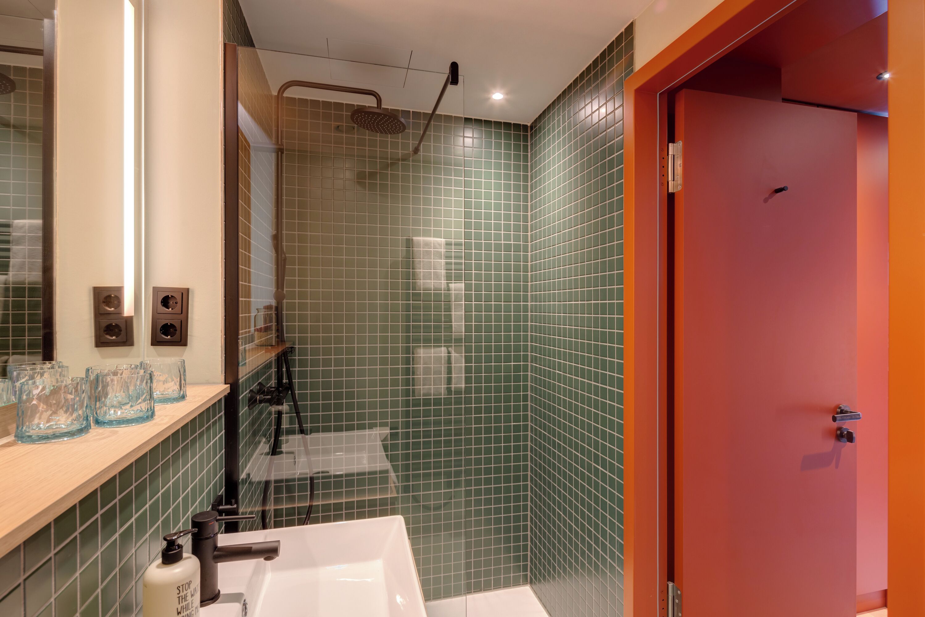 Une salle de bain moderne avec des murs recouverts de carreaux verts, un lavabo blanc et une porte rouge.
