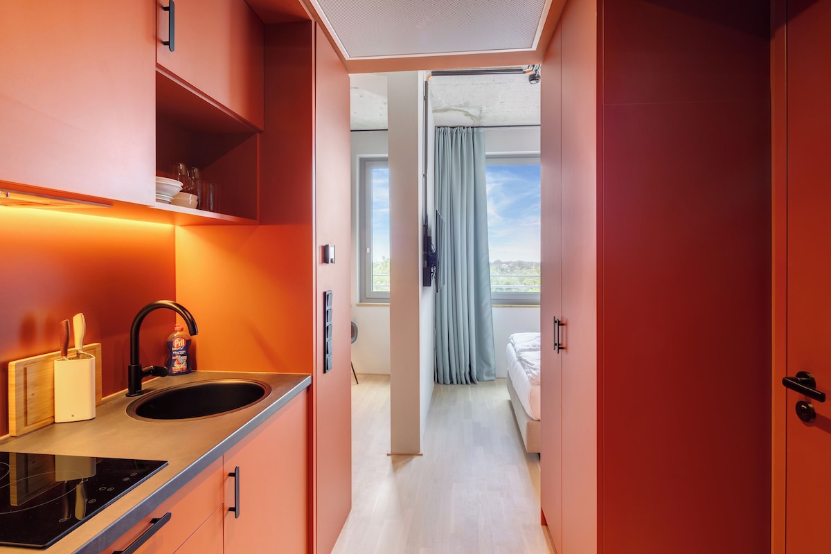 A modern kitchen with a black sink, a built-in stove, and a wooden utensil holder. A window with curtains leads to a bedroom with a bed and a view of the outdoors.