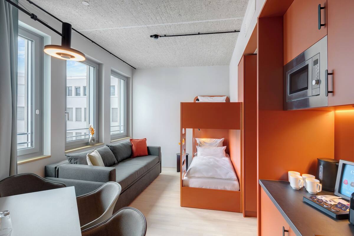 A modern hotel room with a sofa, a bed with a built-in desk, a microwave, and a coffee machine.