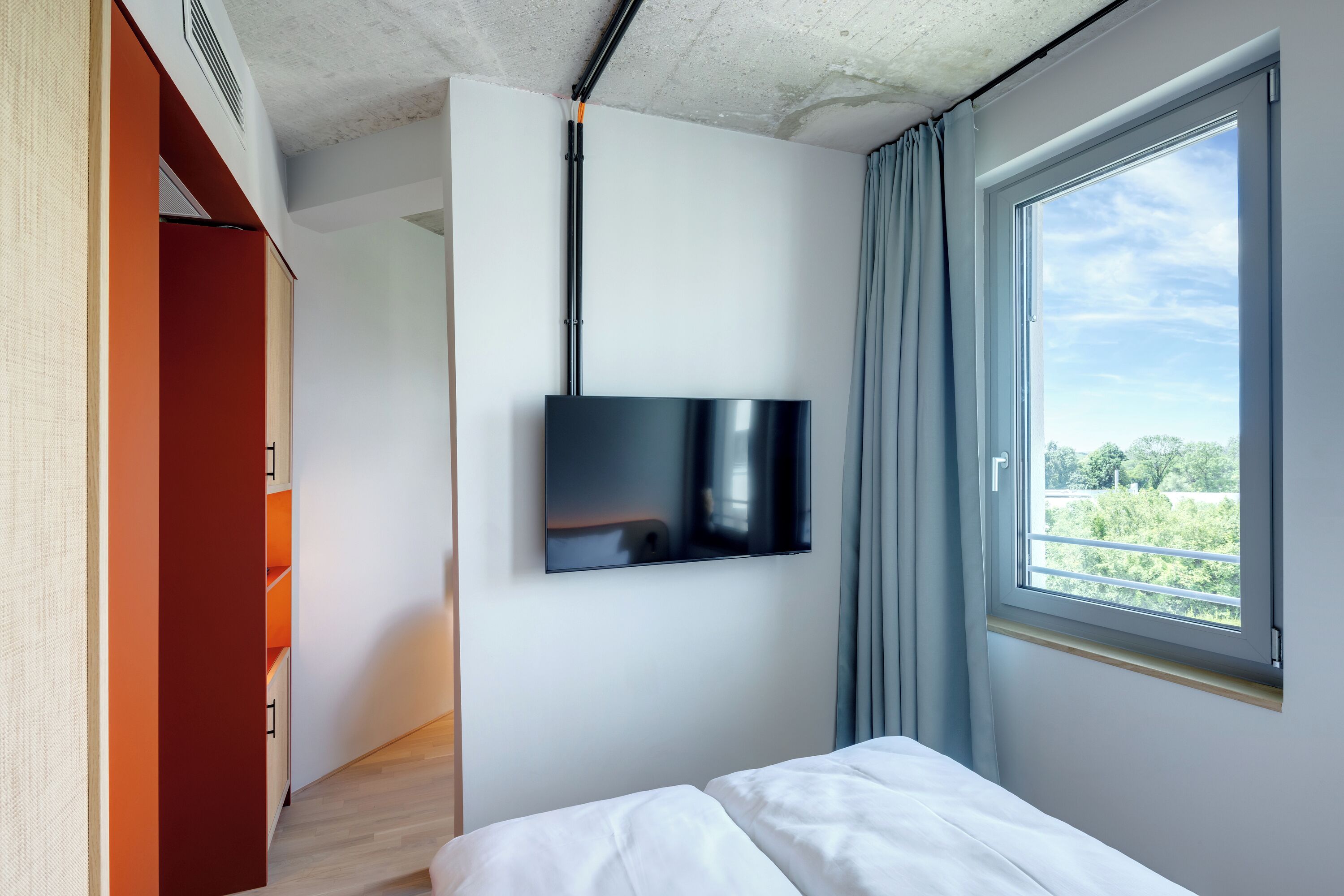 A modern hotel room with a large window, a flat-screen TV mounted on the wall, and a wooden floor.