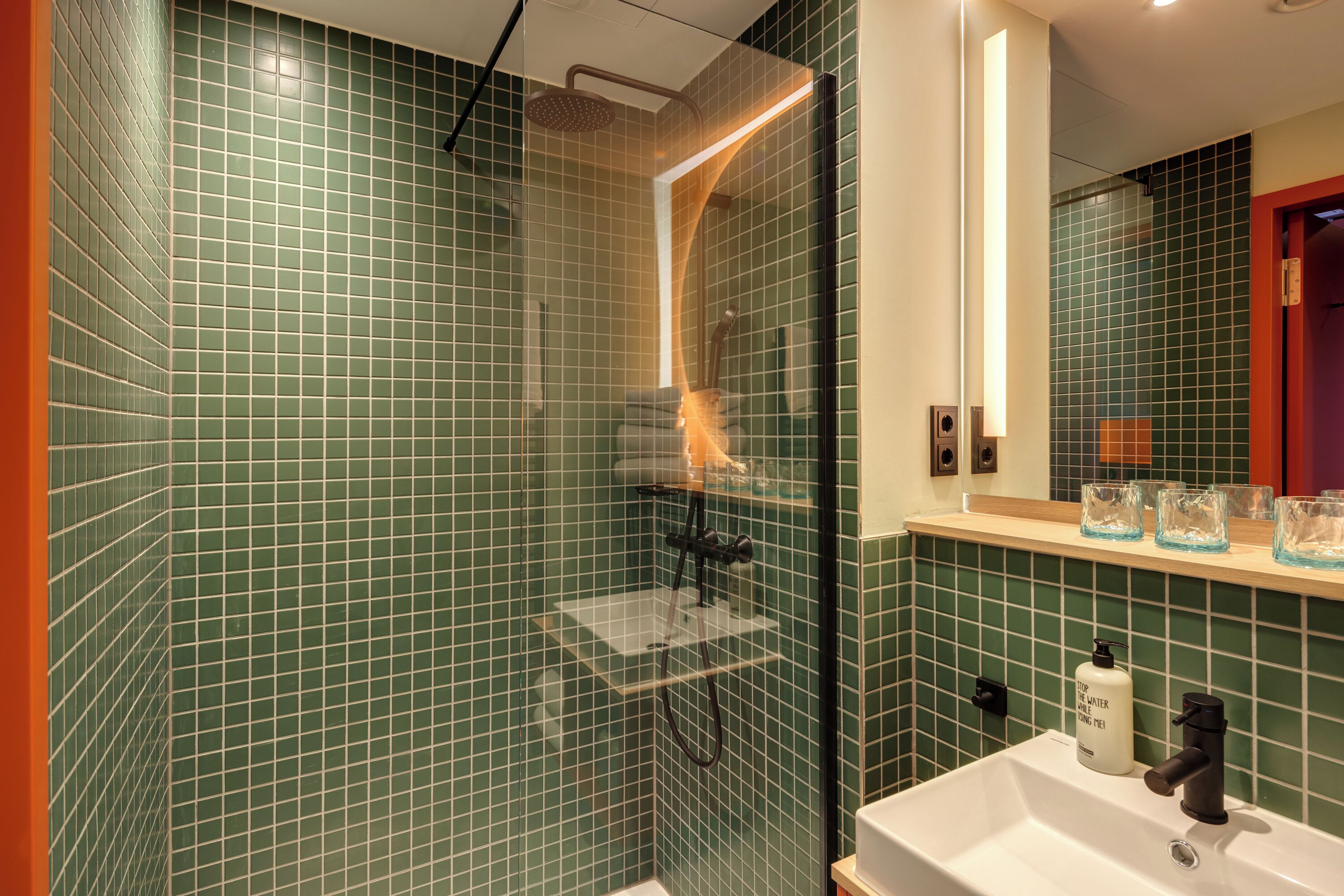A bathroom with green tiled walls, a glass-enclosed shower, a white sink, and a mirror above it.