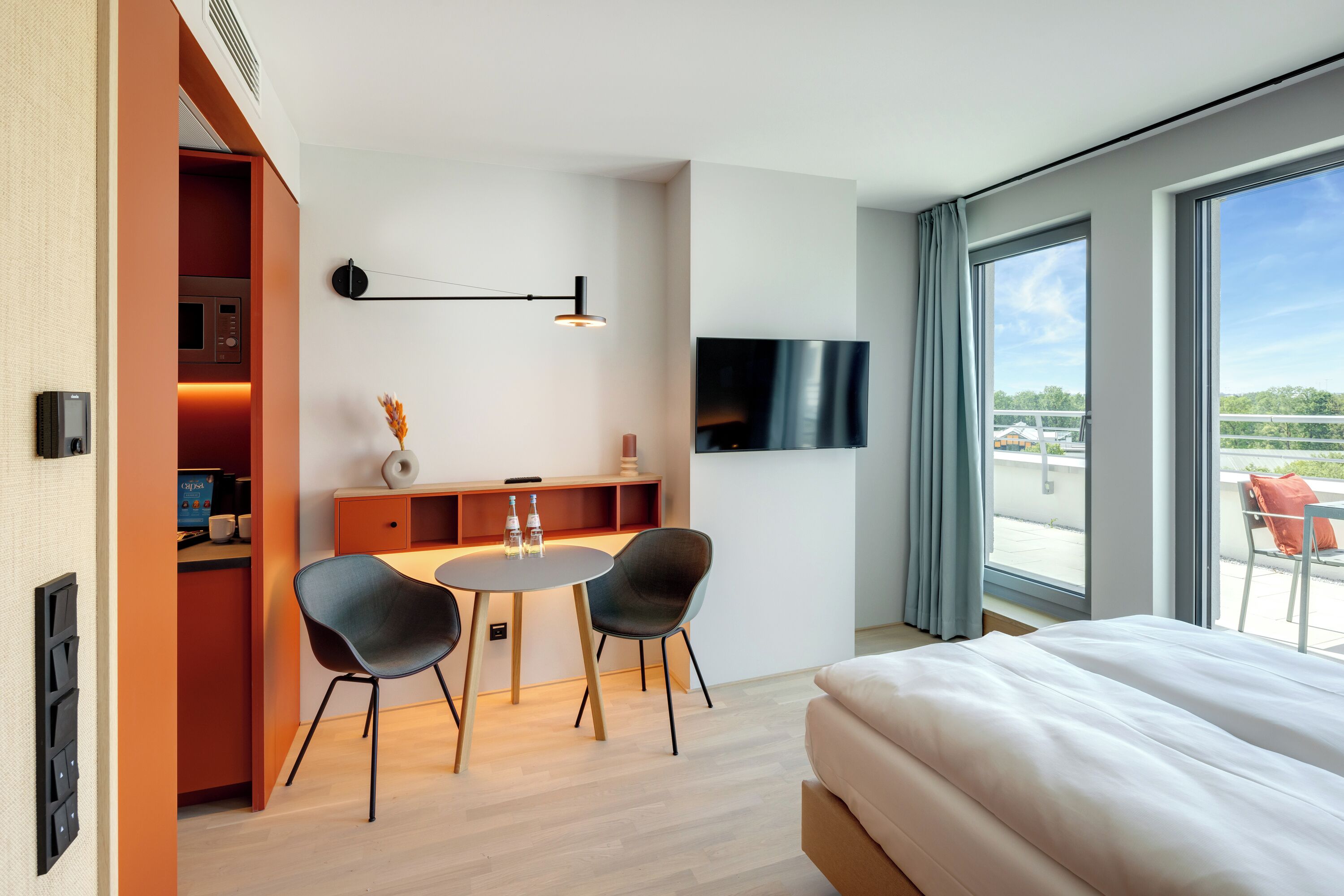 Una habitación de hotel moderna con una cama, un escritorio con computadora, dos sillas, un televisor y un balcón con vistas.
