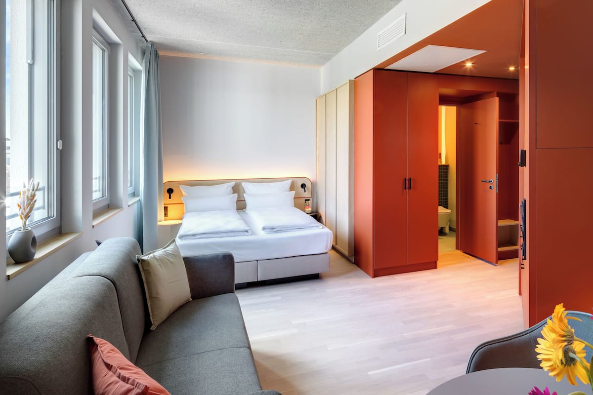A modern hotel room with a bed, a sofa, and a bathroom visible through an open door.