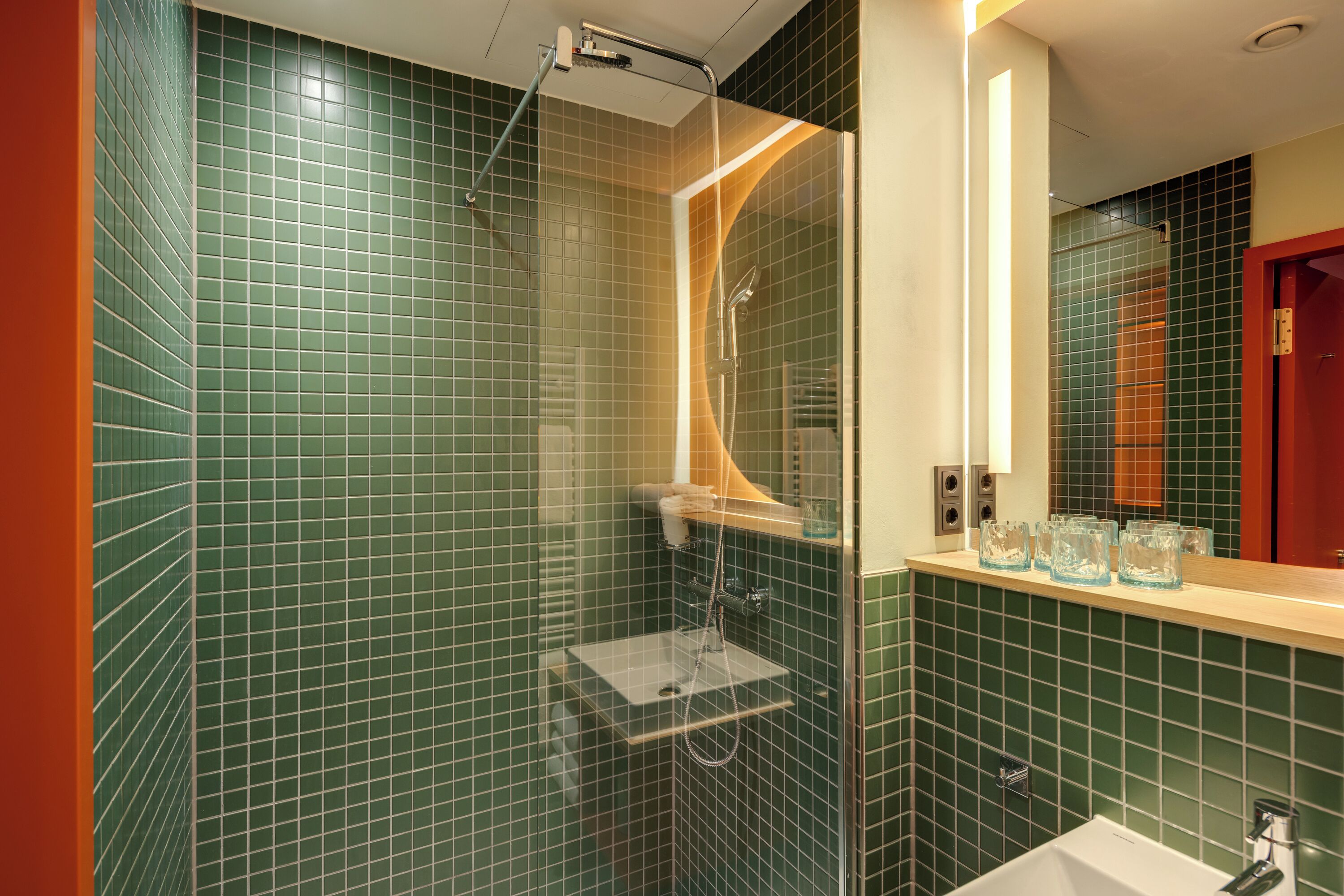 A bathroom with green tiled walls, a glass shower enclosure, a white sink, and a mirror above the sink.